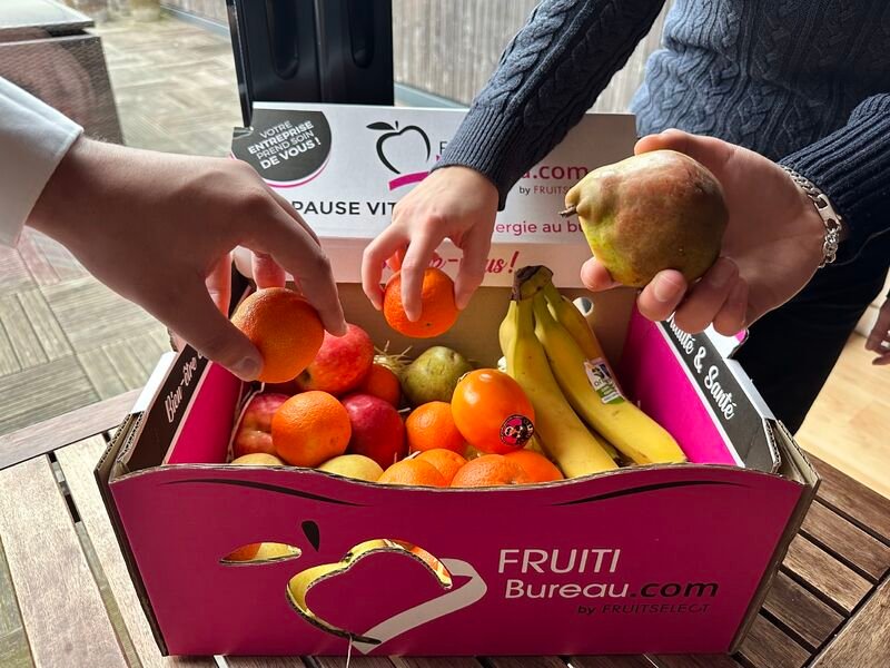 Des mains saisissent clémentines, bananes et poire dans une caisse Fruitibureau posée sur une table en bois